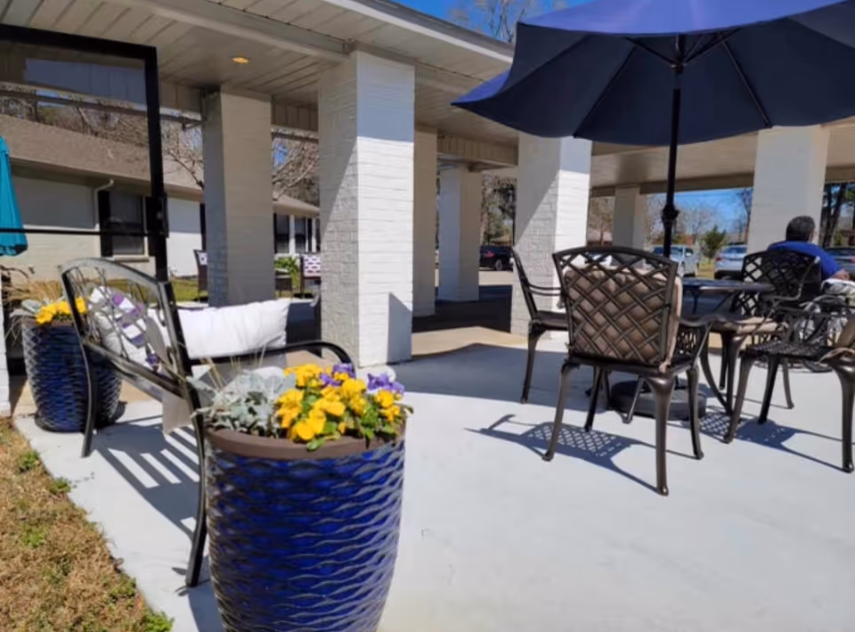 Outdoor patio area at Magnolia Place Wellness and Rehabilitation with metal chairs and tables under a large blue umbrella. There are large blue planters with yellow and purple flowers, white brick pillars, and a person sitting at one of the tables. The sky is clear and blue.
