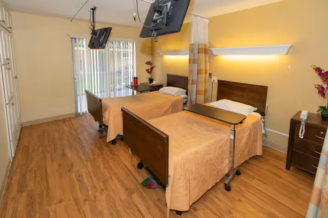 A healthcare center room with two hospital beds covered in beige bedspreads, each equipped with an overbed table. The room has wood flooring, a sliding glass door with vertical blinds, two wall-mounted televisions above the beds, and bedside tables with a telephone and flowers. A privacy curtain is partially drawn between the beds.