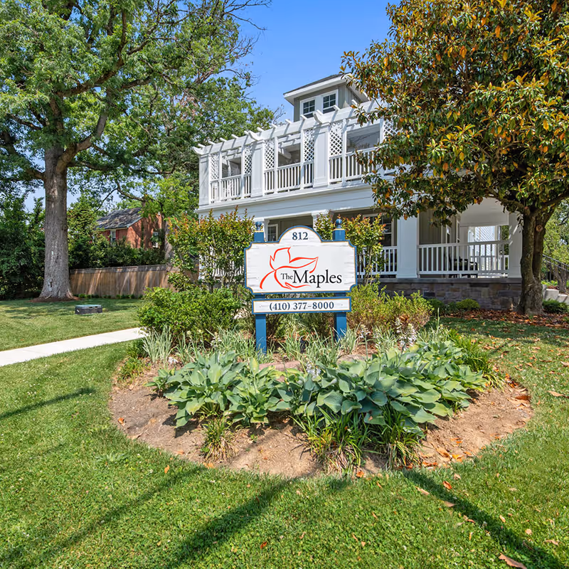 Exterior view of a white two-story building with a porch and latticework on the upper balcony, surrounded by trees and greenery. In front of the building is a landscaped garden bed with plants and a sign that reads 'The Maples' along with the phone number (410) 377-8000.