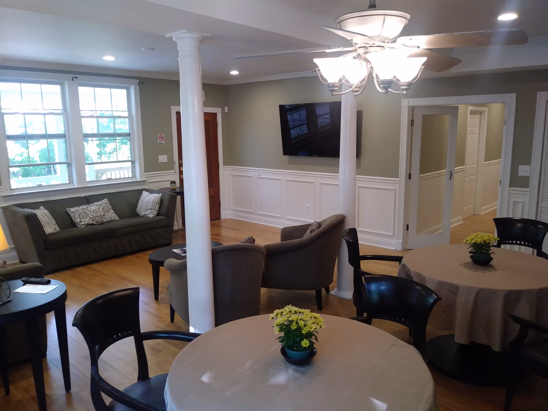 A cozy living room area with wooden floors, two round tables covered with beige tablecloths and small potted yellow flowers on top. There are black chairs around the tables. In the background, there is a gray sofa with patterned cushions, two gray armchairs, a wall-mounted flat-screen TV, and large windows letting in natural light. White columns and a ceiling fan with lights are also visible.
