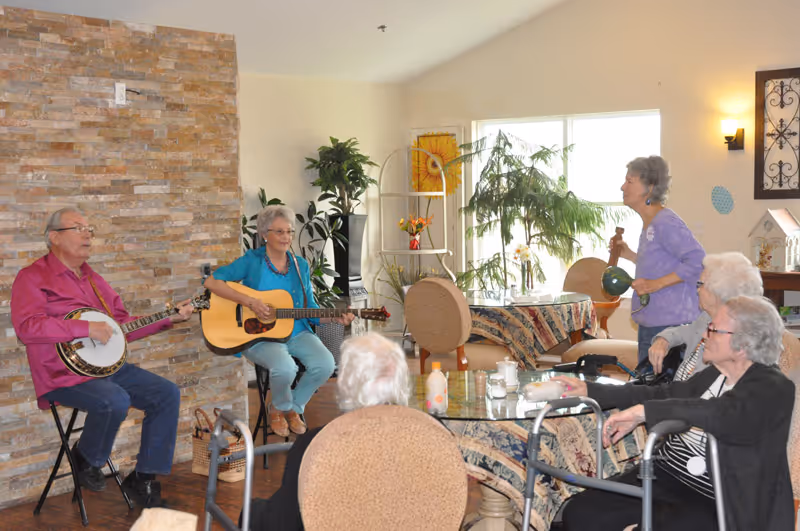 A group of elderly people in a common room at an assisted living facility. Two individuals are playing musical instruments, one with a banjo and the other with a guitar, while another person stands playing a percussion instrument. Several seated residents watch and listen, some using walkers. The room is decorated with plants, a stone accent wall, and large windows letting in natural light.