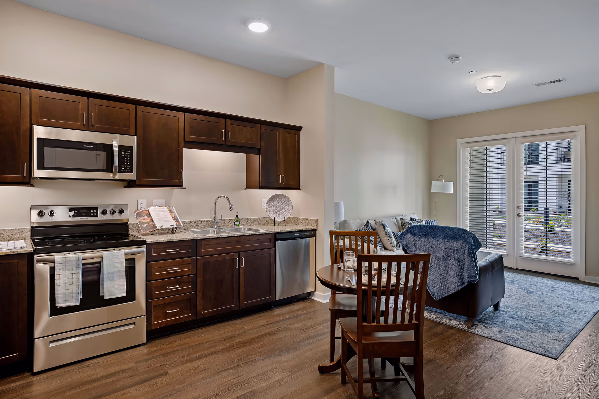 Open kitchen with dark wood cabinets and stainless-steel appliances opening to a small dining area and living room with a sofa and glass doors.