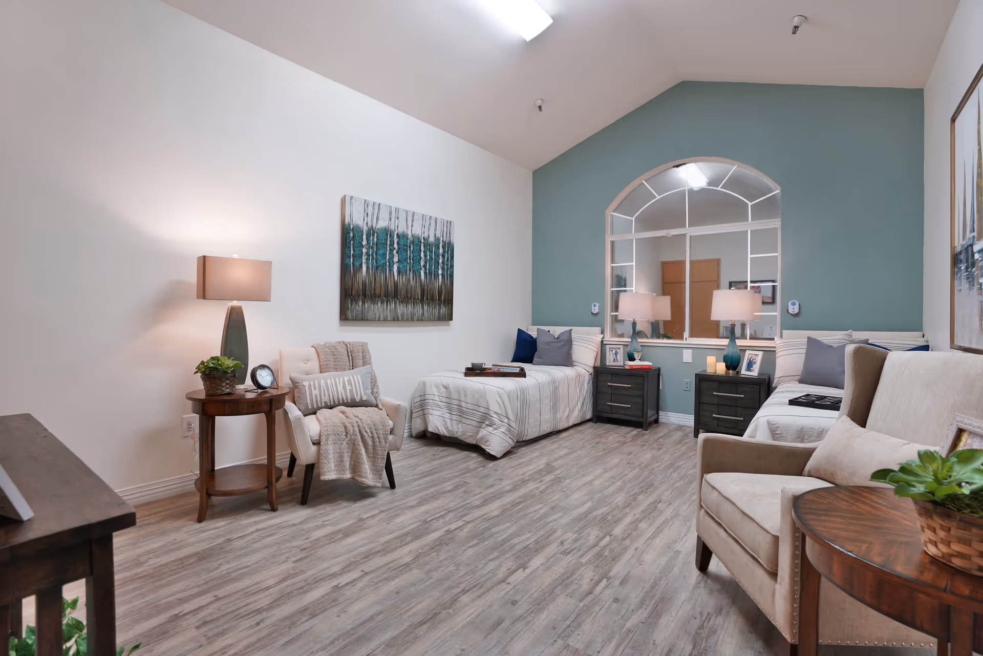 A cozy memory care bedroom with two single beds, each with striped bedding and pillows, separated by two dark nightstands with matching lamps. The room features a large arched window with a mirror, light wood flooring, a beige armchair with a 'THANKFUL' pillow and a throw blanket, a small round wooden side table with a plant and clock, and calming wall art. The walls are painted white and teal.