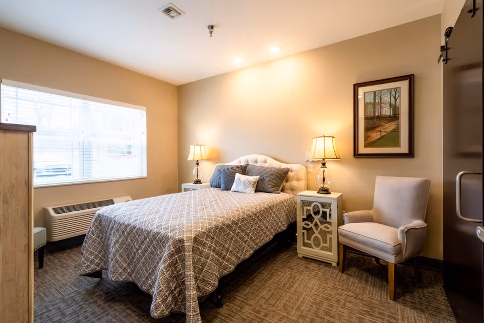 A cozy bedroom with a double bed covered in a patterned gray bedspread and several pillows. There are two bedside tables with lamps on each side of the bed. A cushioned armchair is placed near the right bedside table. A framed picture hangs on the wall above the armchair. A large window with blinds lets in natural light, and a heating/cooling unit is below the window. The room has beige walls and carpeted flooring.