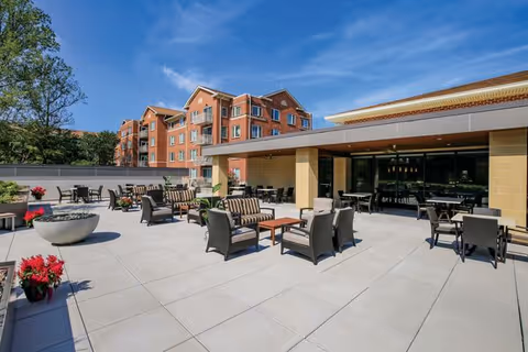 Outdoor patio area with multiple seating arrangements including cushioned chairs and tables, potted plants with red flowers, and a large fire pit. In the background, there is a multi-story brick building under a clear blue sky.