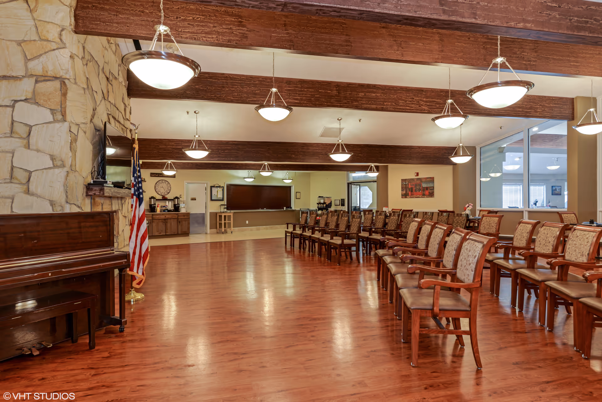 A spacious room with wooden floors and exposed wooden beams on the ceiling, featuring rows of wooden chairs with cushioned seats and backs arranged facing a large flat-screen TV mounted on a stone wall. An American flag stands next to a piano on the left side of the room. The room is well-lit with multiple hanging light fixtures and has a counter area with coffee machines and a clock in the background.