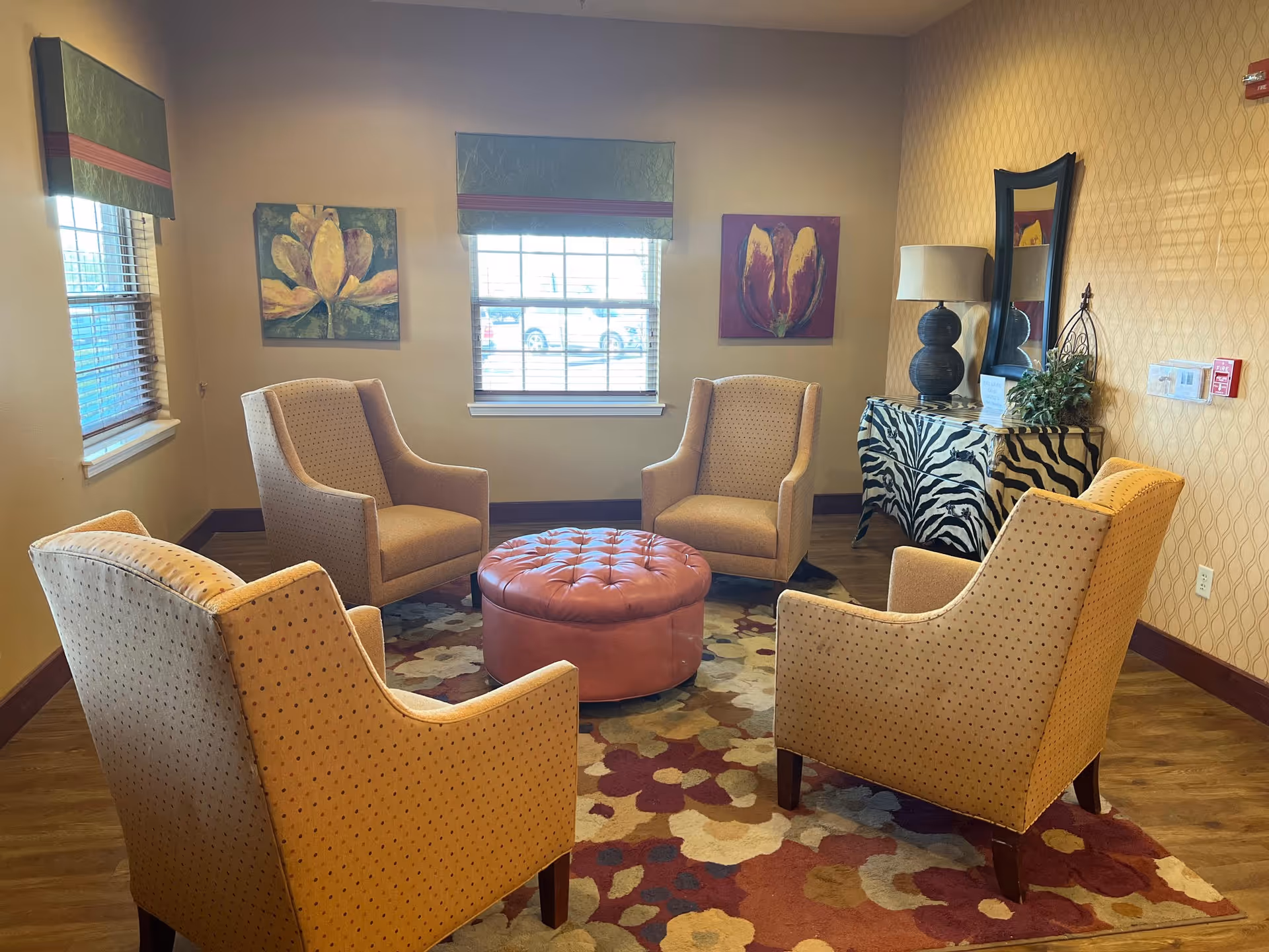 A cozy common sitting area with four upholstered armchairs arranged around a round tufted ottoman on a patterned rug, with windows, artwork, and a zebra-print sideboard.