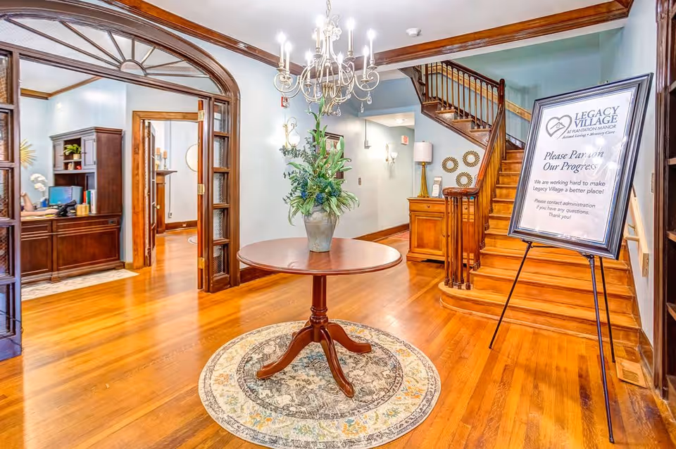 Interior view of a senior living facility lobby with polished wooden floors, a round wooden table with a floral arrangement in the center on a circular rug, a chandelier hanging from the ceiling, a wooden staircase leading upstairs, and a sign on an easel that reads 'Legacy Village at Plantation Manor Please Pardon Our Progress'.