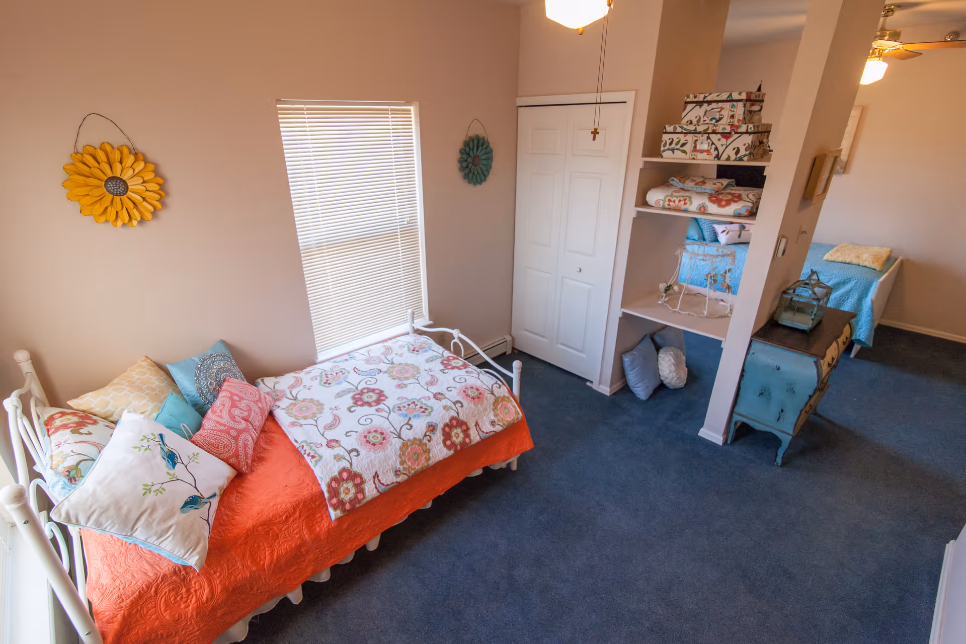A cozy bedroom with two single beds, each decorated with colorful pillows and floral-patterned quilts. The room has a window with blinds, a closet with white double doors, and built-in shelves holding decorative boxes and pillows. The carpet is blue, and the walls are painted a soft beige. A yellow flower wall decoration hangs near the window.
