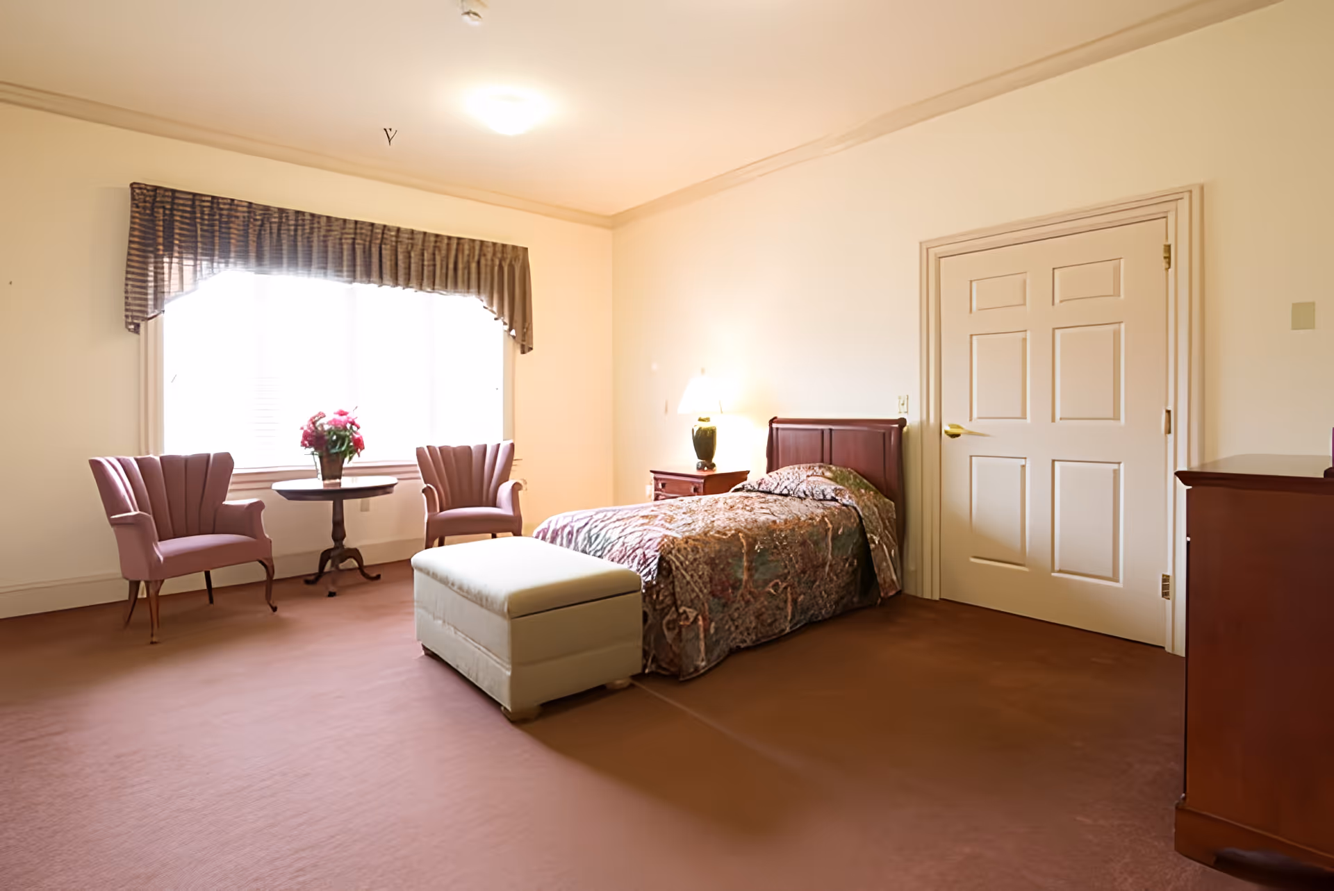 A cozy bedroom with a single bed covered in a patterned bedspread, a small nightstand with a lamp, two pink upholstered chairs with a round wooden table between them holding a vase of flowers, a window with a valance curtain, a beige ottoman at the foot of the bed, and a closed white door.