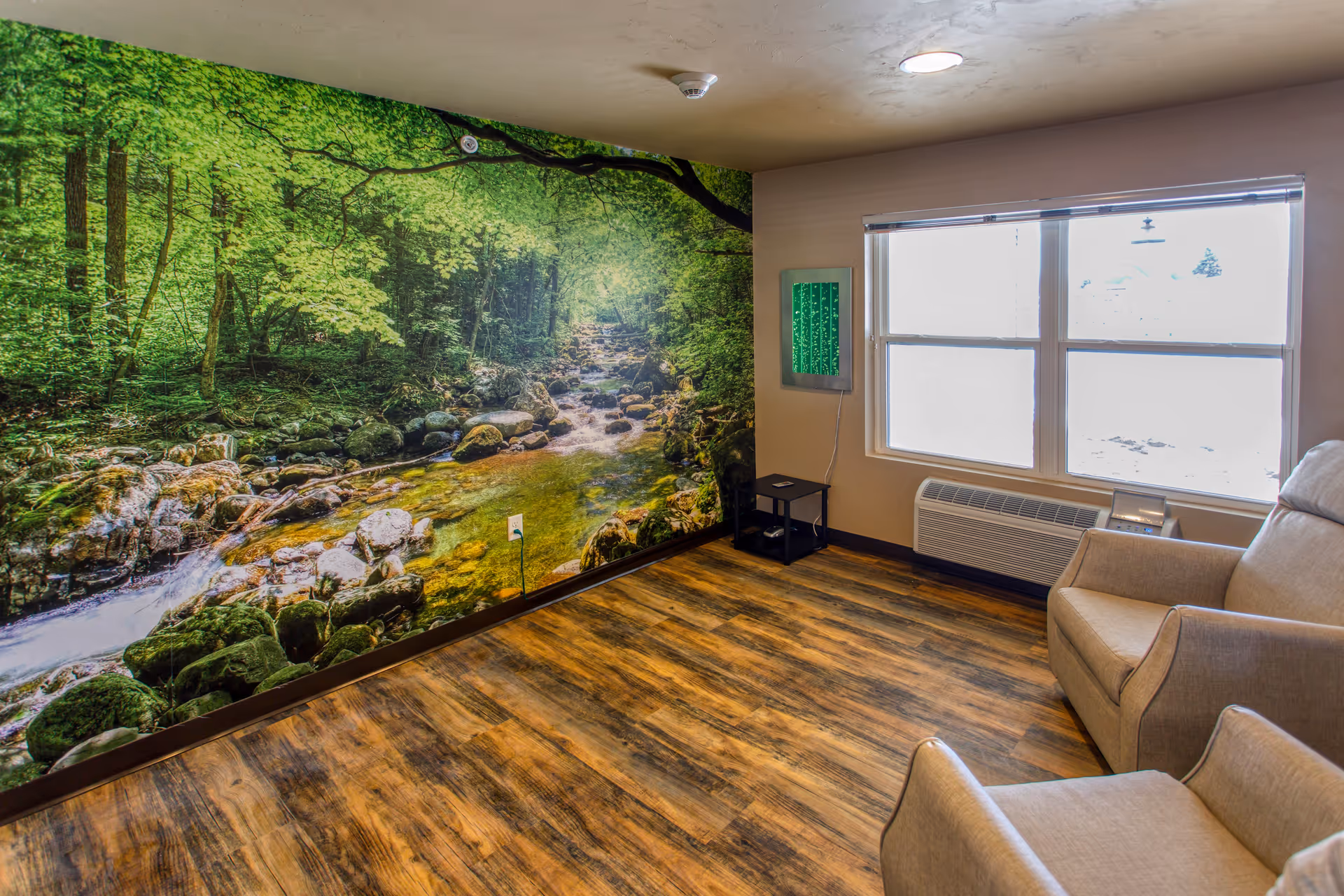 A cozy room with two beige armchairs, a large window with blinds, and a wall mural depicting a serene forest stream with rocks and green trees. The floor has wood-like vinyl flooring, and there is a small black side table with a green light panel mounted on the wall above it.