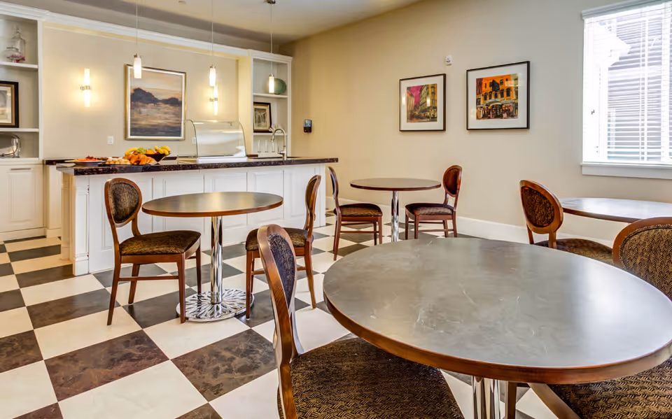 A bright dining area with round tables and wooden chairs with cushioned seats. The floor has a black and white checkered pattern. In the background, there is a counter with a fruit bowl and a covered food display. The walls are decorated with framed artwork and there is a window with blinds letting in natural light.