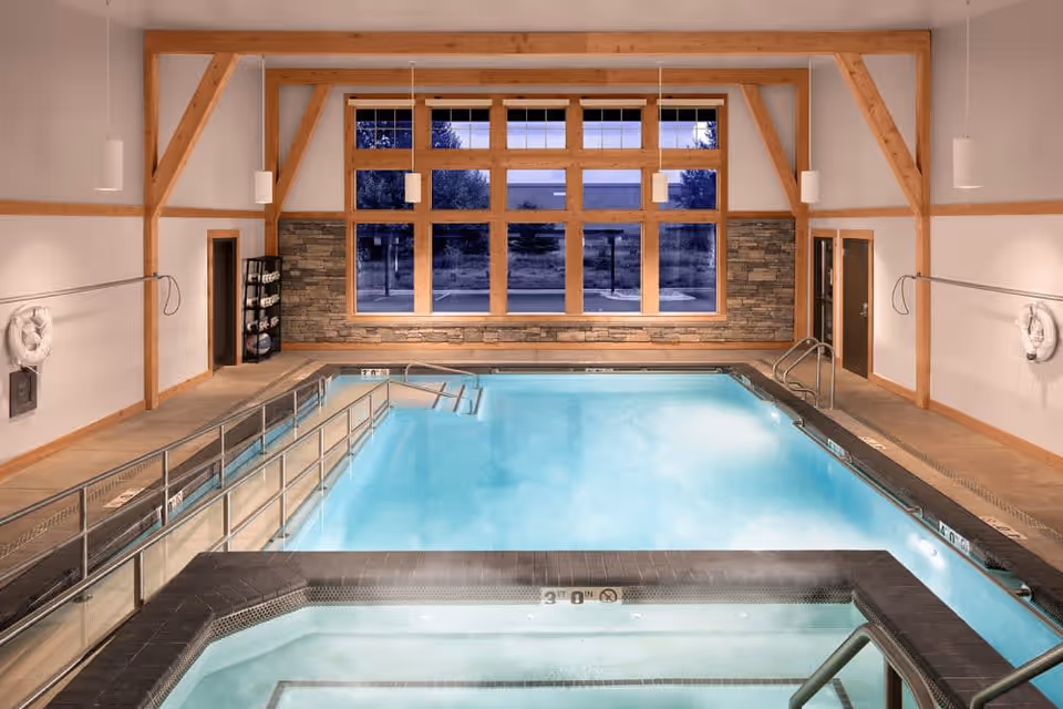 Indoor swimming pool area with a smaller hot tub in the foreground, large windows with wooden frames letting in natural light, stone accent wall, wooden beams on the ceiling, and safety equipment on the walls.