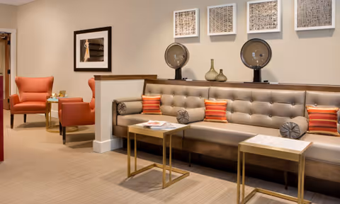 A stylish senior living lounge with tufted bench seating, two small marble-top tables, orange armchairs, and wall art.