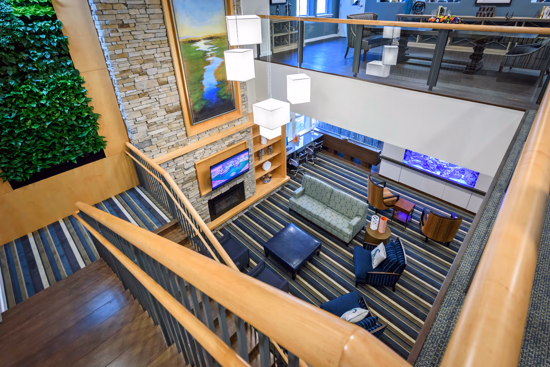 View from the top of a staircase looking down into a modern living room area with striped carpet, a green sofa, two blue armchairs, a black ottoman, and a small round table. The room features a stone fireplace with a mounted TV above it, a large vertical painting, hanging cube-shaped pendant lights, a wall with green plants, and a fish tank built into the wall. There is a glass railing on the upper level overlooking the living area.