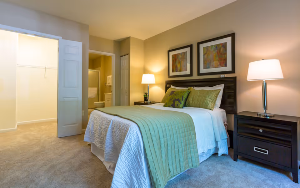 Neatly made bedroom with a bed dressed in green bedding, two bedside tables with lamps, artwork above the headboard, and an open doorway leading to a closet and bathroom.