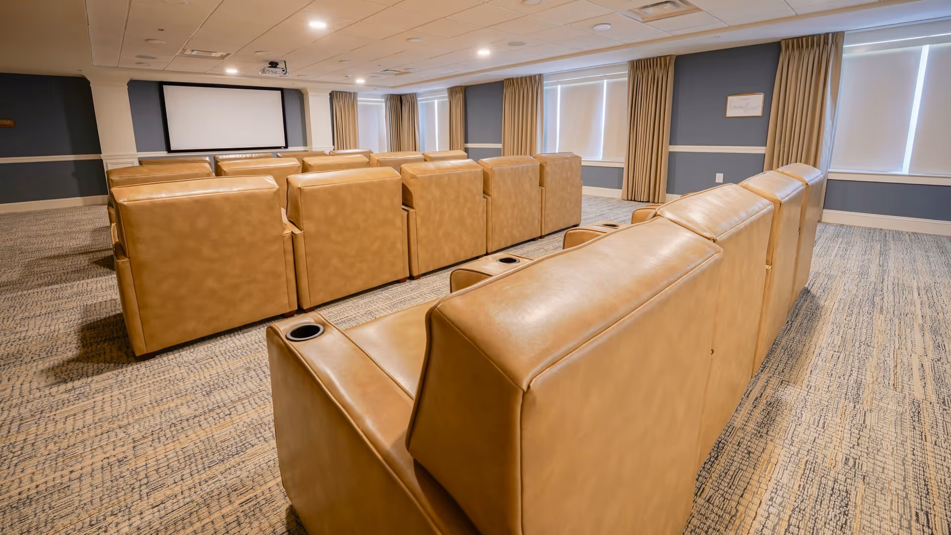 A cozy media room with multiple rows of tan leather recliner chairs facing a large projector screen. The room has carpeted flooring, blue-gray walls with white trim, and windows with beige curtains allowing natural light.