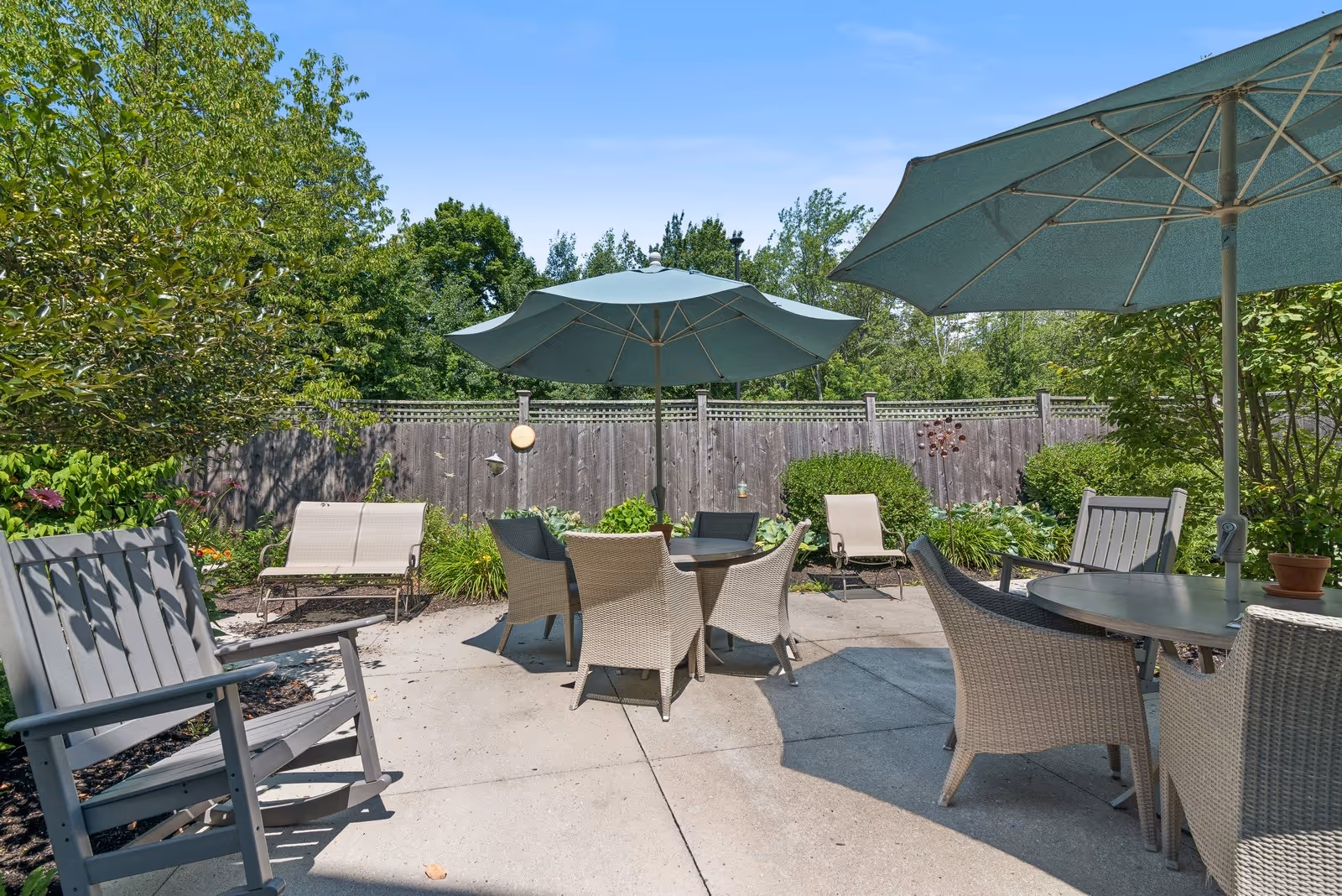Outdoor patio area with several tables and chairs under large green umbrellas, surrounded by greenery and a wooden fence under a clear blue sky.