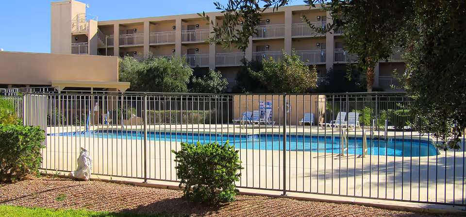 Outdoor swimming pool area enclosed by a metal fence with lounge chairs around the pool. In the background, there is a multi-story building with balconies and some trees and bushes surrounding the pool area.