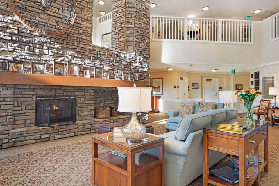 A cozy living room area with a large stone fireplace, a light blue sofa with patterned cushions, wooden side tables with lamps, and a vase of flowers. The room has a high ceiling with a balcony railing above, framed pictures on the fireplace mantel, and warm lighting.