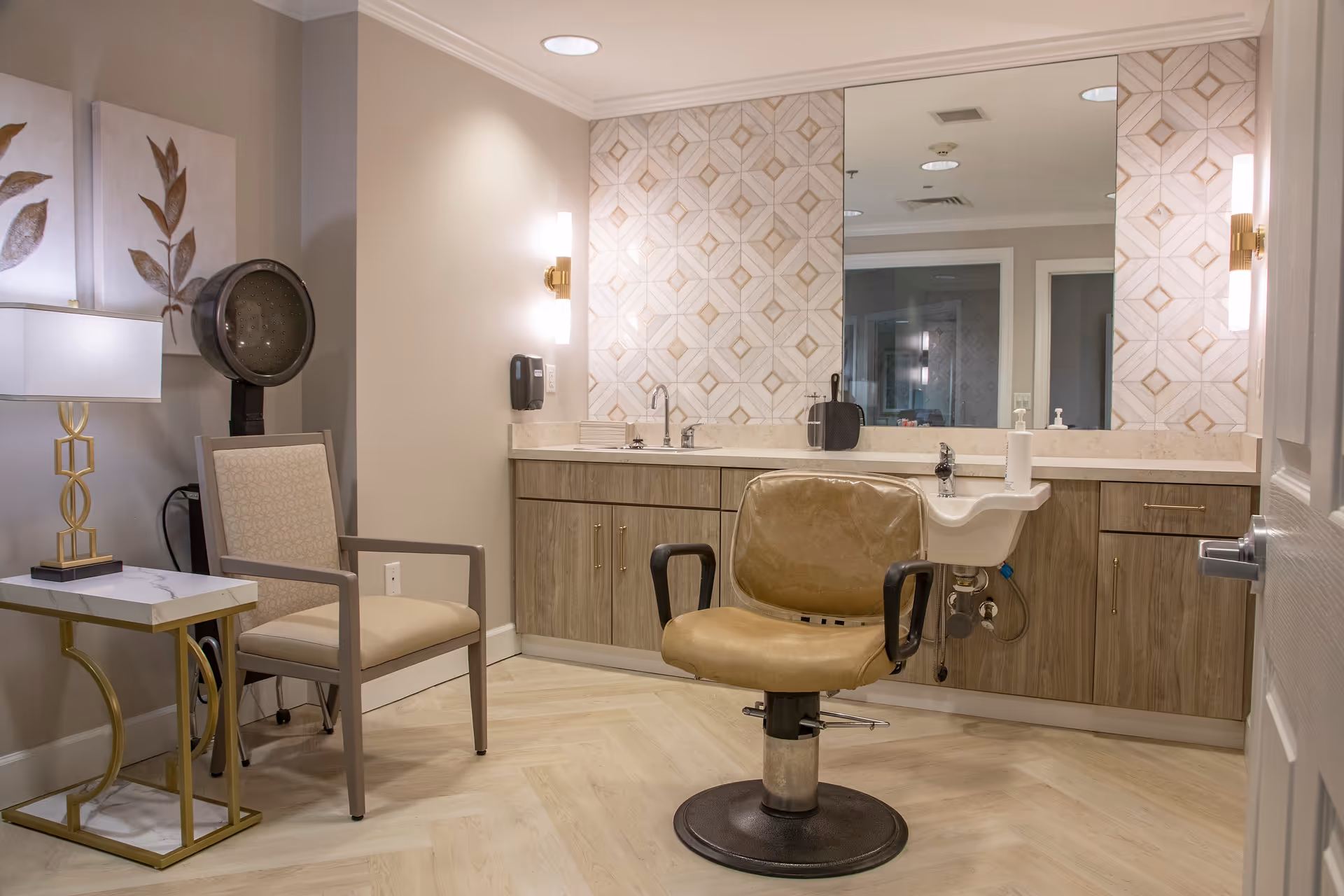 Interior of a hair salon or grooming area with a beige salon chair in front of a large mirror and a sink. There is a beige armchair next to a small table with a lamp, and a hair dryer hood in the corner. The walls are light-colored with decorative tiles around the mirror.