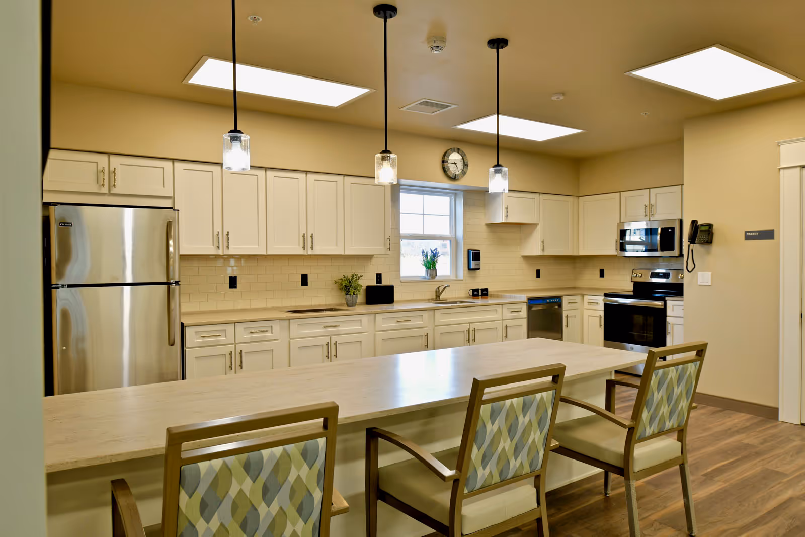 A modern kitchen in a senior living facility featuring white cabinets, stainless steel refrigerator, oven, microwave, and dishwasher. There is a long island countertop with three patterned chairs, pendant lights hanging from the ceiling, a window with a small plant on the sill, and a wall clock above the window.