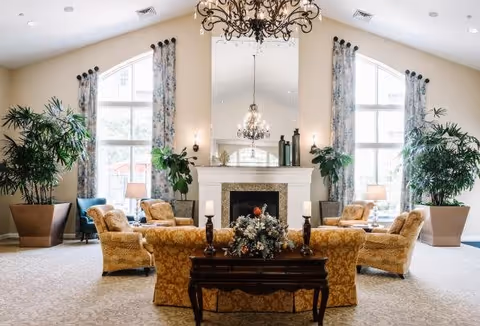 A spacious and elegant living room with a high vaulted ceiling, large arched windows with floral curtains, and a central fireplace with a large mirror above it. The room is furnished with patterned yellow armchairs and sofas arranged around a dark wooden coffee table decorated with candles and a floral centerpiece. There are large potted plants on either side of the fireplace and table lamps providing warm lighting.