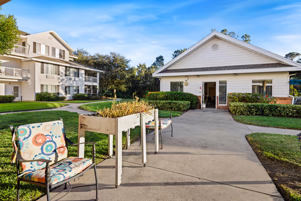Outdoor view of a senior living community showing a paved walkway leading to a white building with a gabled roof. There are two colorful cushioned chairs and a raised garden planter along the walkway. The area is surrounded by green grass, bushes, and trees under a clear blue sky.