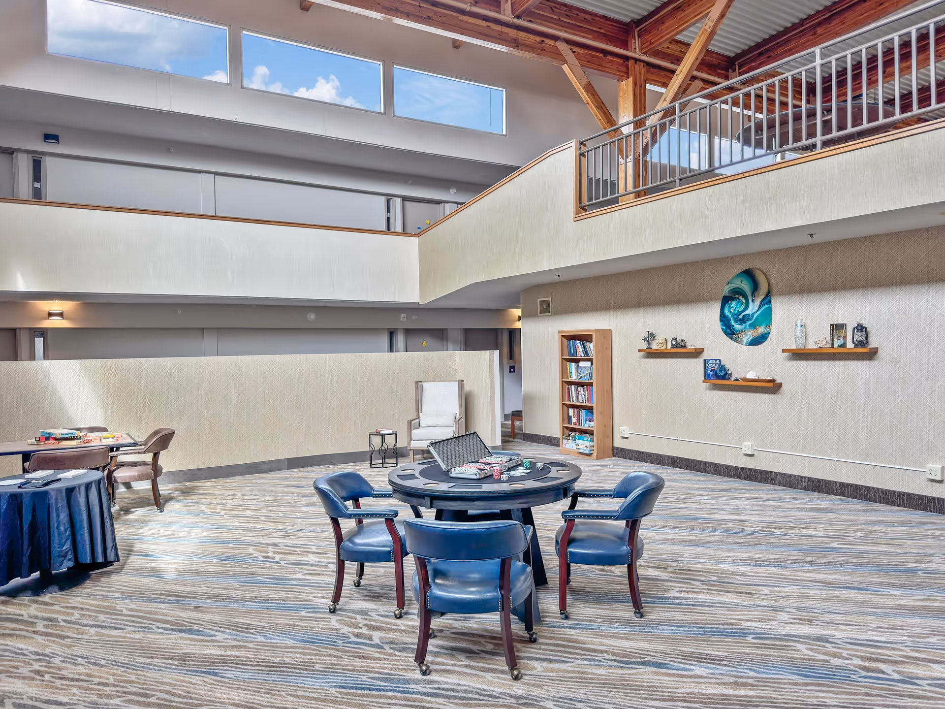 Bright two-story common area with a round game table surrounded by chairs, bookshelves, seating, and an upper balcony with skylights.