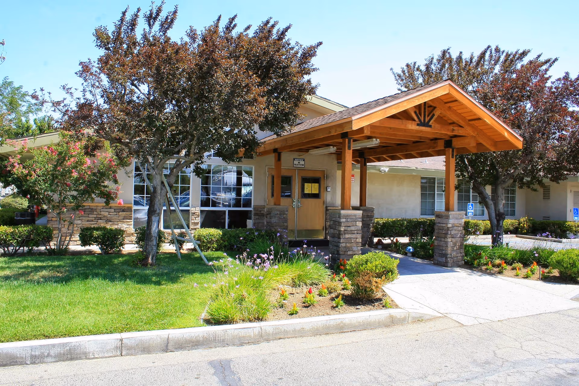 Exterior view of Lancaster Healthcare Center showing the entrance with a wooden canopy supported by stone pillars. The building is surrounded by green grass, bushes, and trees under a clear blue sky.