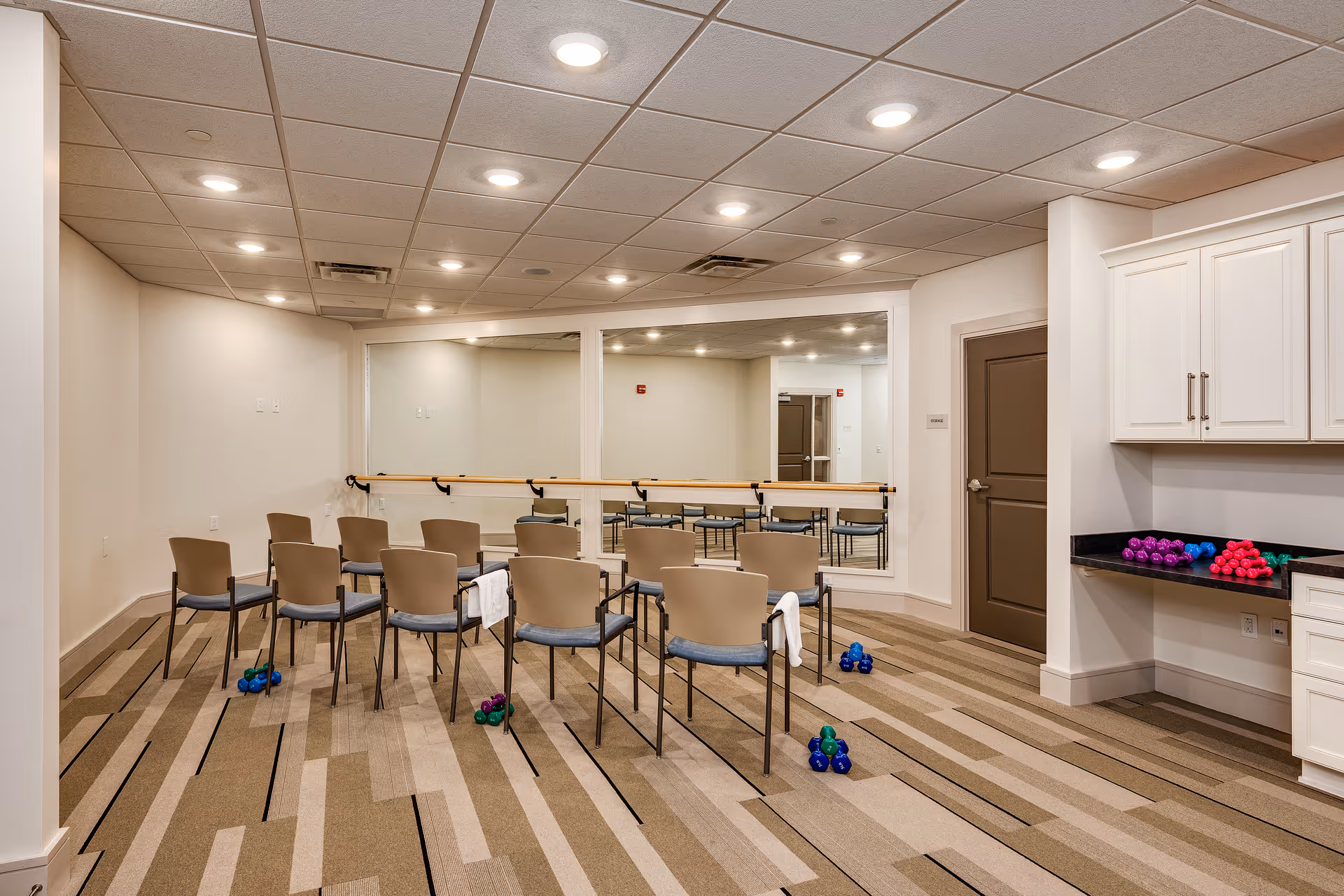 A fitness or exercise room with chairs arranged in rows facing a large mirror on the wall. There are small dumbbells of various colors placed on the floor and on a countertop to the right. The room has a patterned carpet, white walls, a drop ceiling with recessed lighting, and a closed brown door.