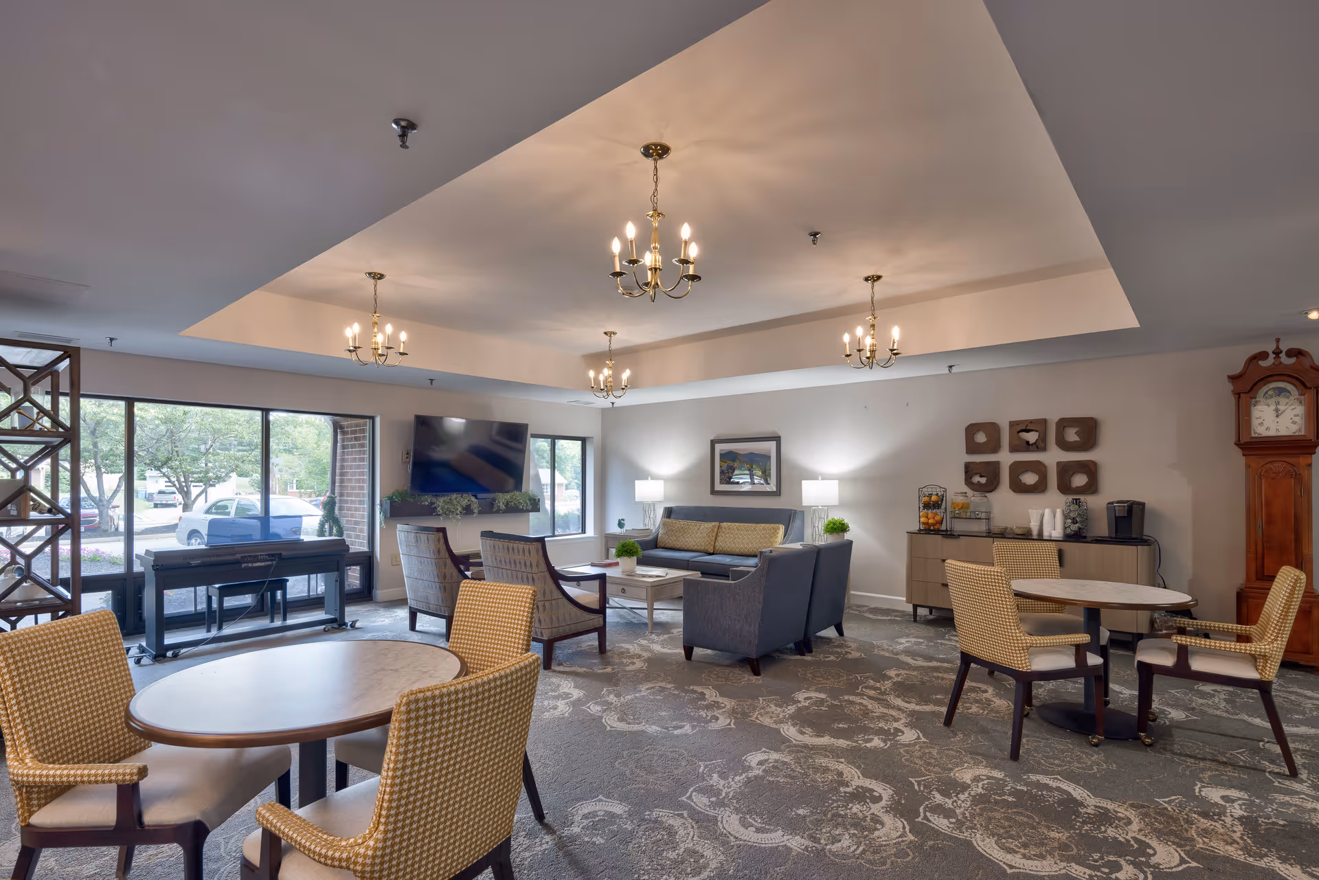 A spacious and well-lit common area with multiple seating arrangements including round tables with chairs and a sofa set with armchairs. The room features large windows letting in natural light, a wall-mounted TV, chandeliers hanging from the ceiling, a piano near the window, a sideboard with a coffee maker and fruit display, and a tall grandfather clock in the corner.