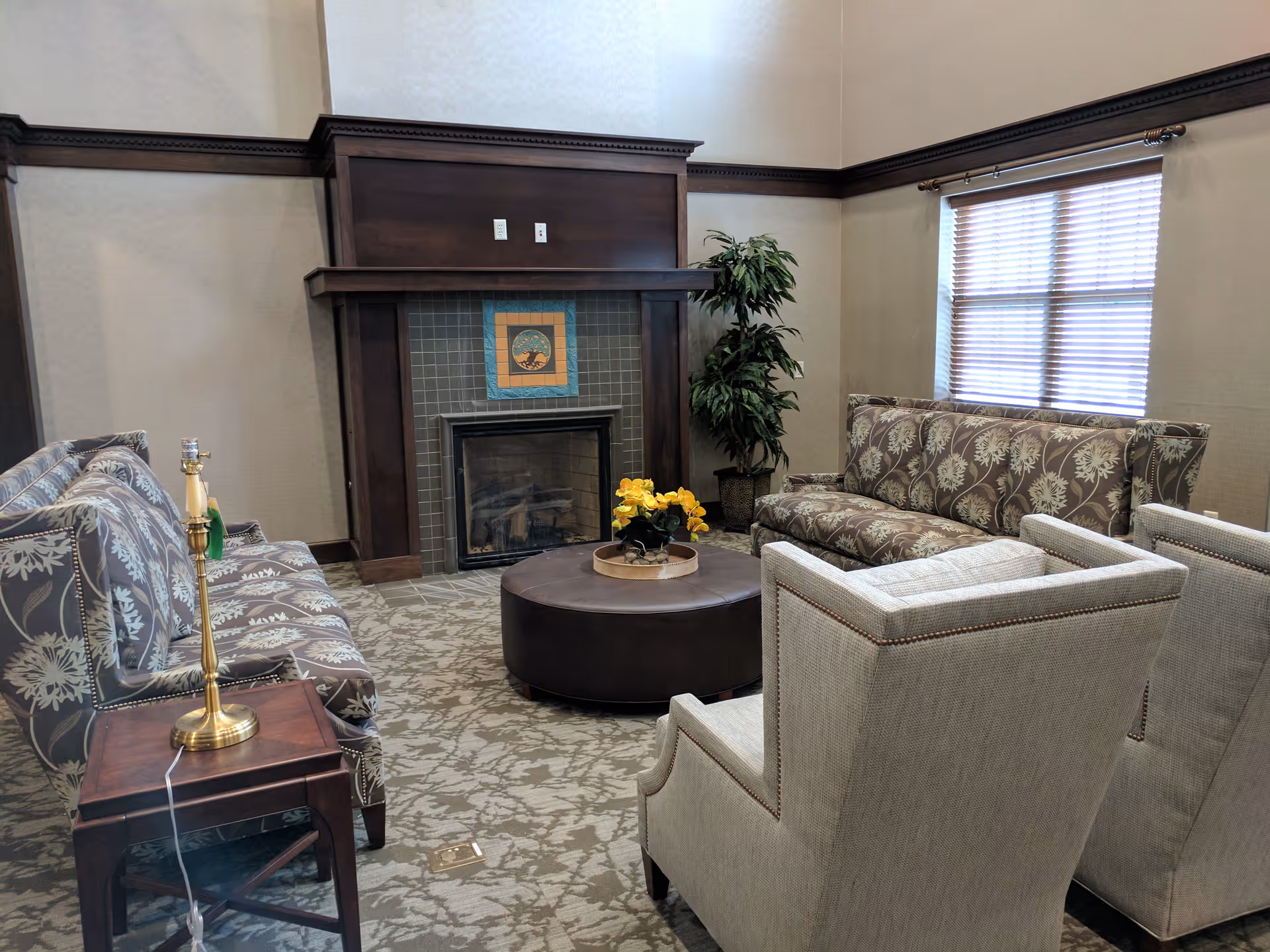 A cozy living room area with patterned sofas and two beige armchairs arranged around a round brown ottoman. There is a fireplace with a decorative tile and wooden mantel, a potted plant in the corner, and a window with blinds letting in natural light.
