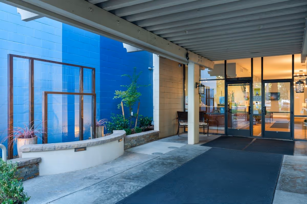Entrance area of Eskaton Land Park facility with a covered walkway, a curved stone bench, potted plants, and glass doors leading inside. The wall on the left is painted in shades of blue, and there is a chair near the entrance.