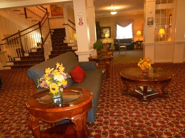 Interior view of a senior living facility lounge area with patterned carpet, a blue sofa with a red pillow, two wooden coffee tables with glass tops and floral arrangements, and a staircase in the background leading to an upper floor.