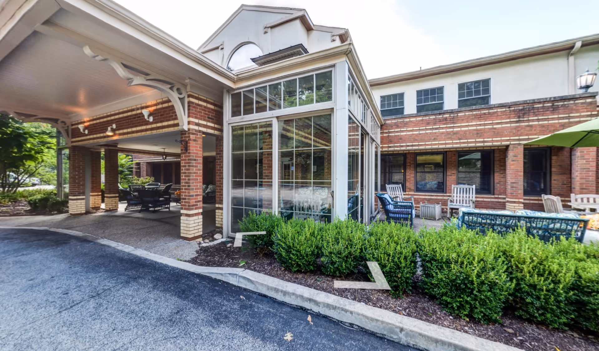 Exterior view of The Pines of Mount Lebanon senior living facility showing a covered entrance with brick pillars, a glass-enclosed sunroom, outdoor seating area with chairs and tables, green bushes, and part of the building with windows.