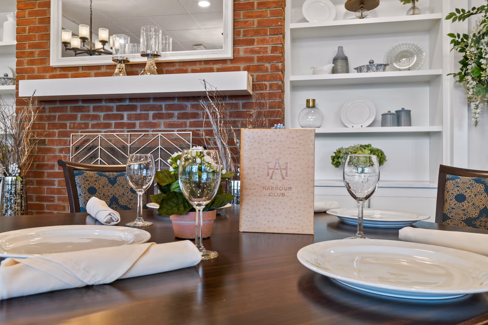 A dining table set with white plates, folded napkins, and empty wine glasses in front of a brick fireplace with a white mantel. A menu labeled 'Harbour Club' and a small potted plant are on the table. Shelves with decorative plates and vases are visible in the background.
