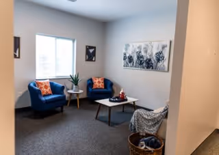 A cozy living room area with two blue armchairs featuring orange patterned pillows, a small round wooden side table with a plant, a white rectangular coffee table with decorative items, a gray armchair with a blanket draped over it, and wall art including a large floral painting and two smaller framed pictures. The room has a window letting in natural light and carpeted flooring.