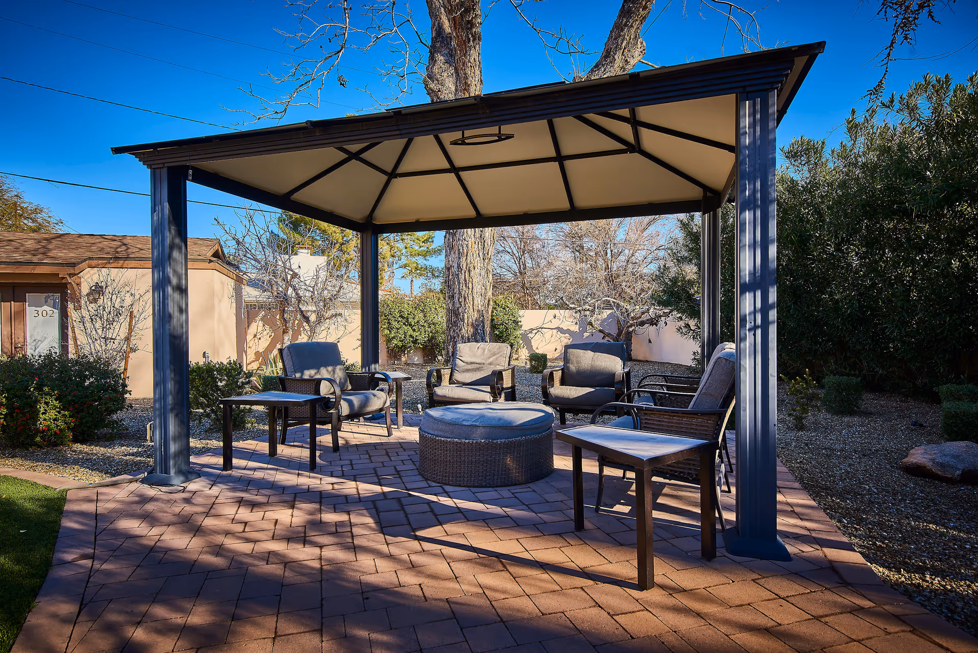 Outdoor seating area under a gazebo with cushioned chairs and tables on a paved patio surrounded by trees and shrubs under a clear blue sky.