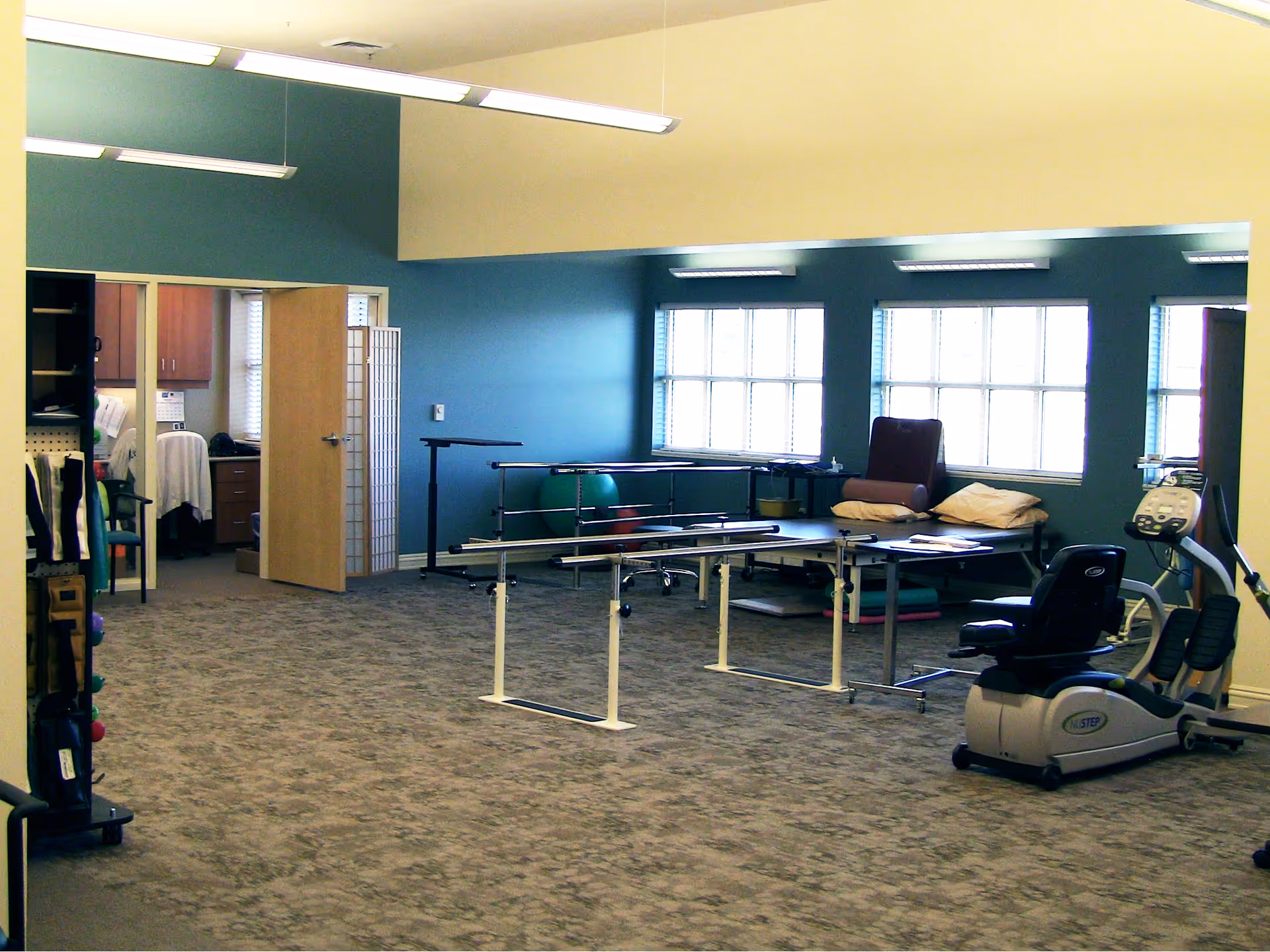 A spacious room with exercise and physical therapy equipment including parallel bars, an exercise bike, and a padded therapy table with pillows. The room has large windows letting in natural light, blue and beige walls, and carpeted flooring. An open door leads to an adjacent office or storage area.