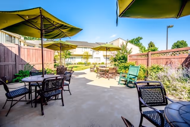 Outdoor patio area with several round tables and chairs under large green umbrellas, surrounded by a wooden fence and greenery, with a building visible in the background under a clear blue sky.