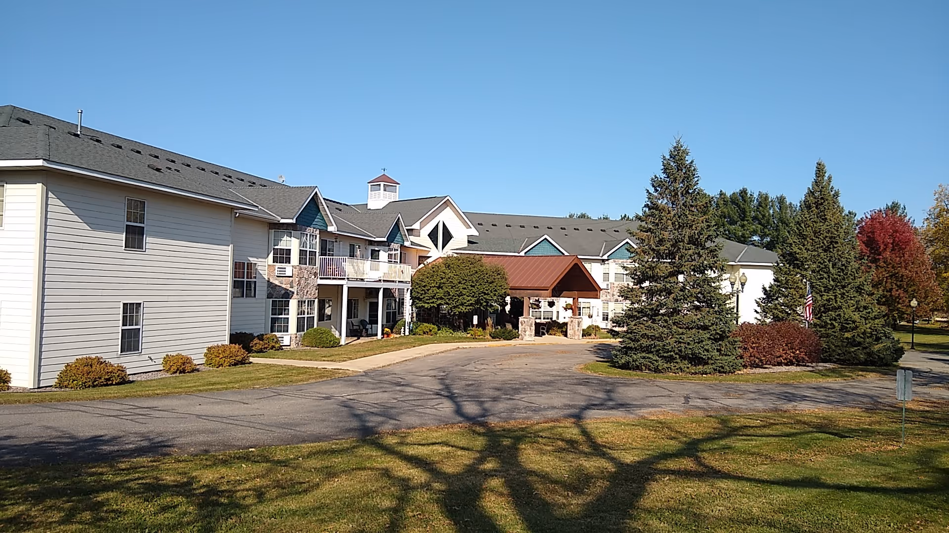 Exterior view of a senior living facility building with white siding and green roof accents, surrounded by trees and shrubs under a clear blue sky. There is a driveway leading to a covered entrance with stone pillars.
