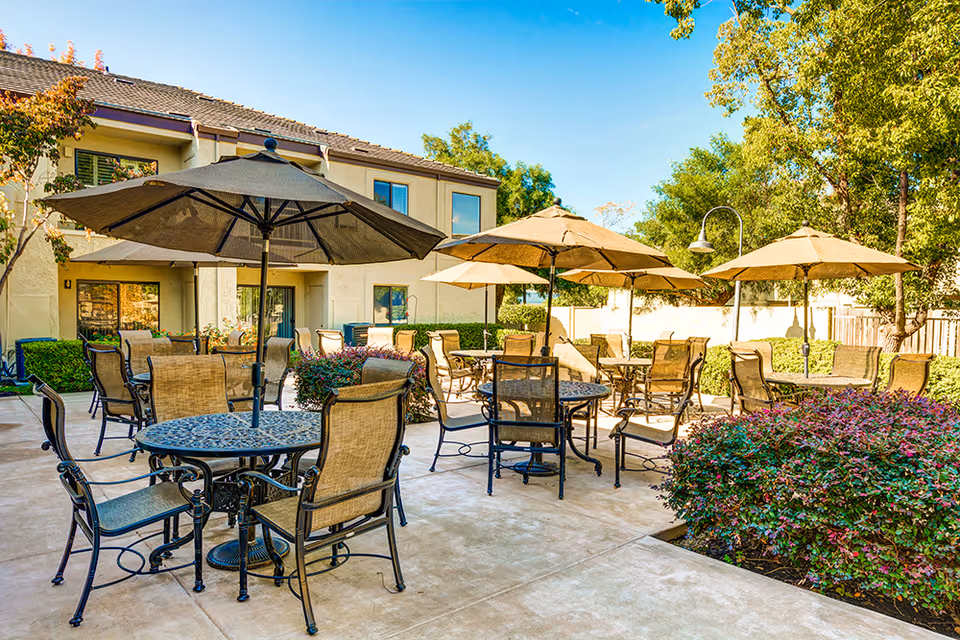 Outdoor courtyard patio with multiple round tables, chairs, and umbrellas in front of a two-story building with landscaping.