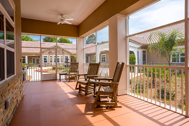 A covered outdoor patio area with several wooden rocking chairs facing outward. The patio is enclosed with white railings and screened windows, overlooking a landscaped courtyard with plants and a building with a tiled roof in the background.