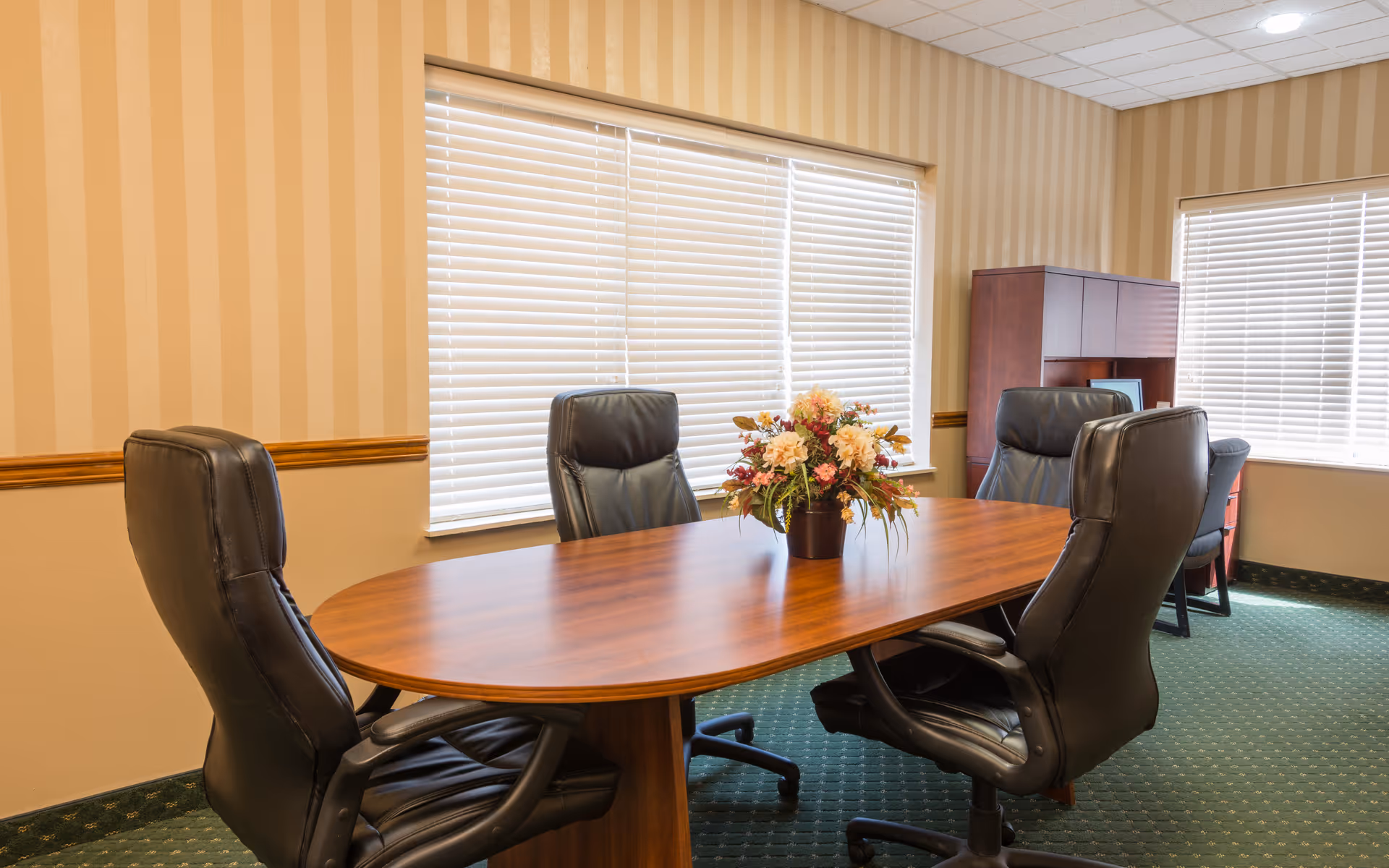 A small conference room with a wooden oval table surrounded by four black leather office chairs. A floral arrangement is placed in the center of the table. The room has beige striped wallpaper, green carpet, two windows with white blinds, and a wooden cabinet with a computer in the corner.