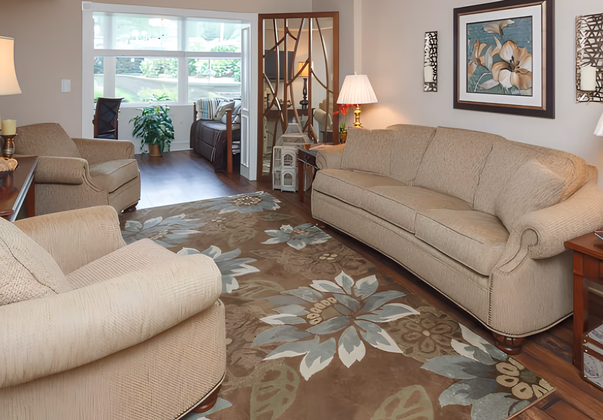 Cozy living room with beige sofa and armchairs arranged around a floral area rug, a side table lamp and framed art, with a windowed sunroom and plants visible in the background.