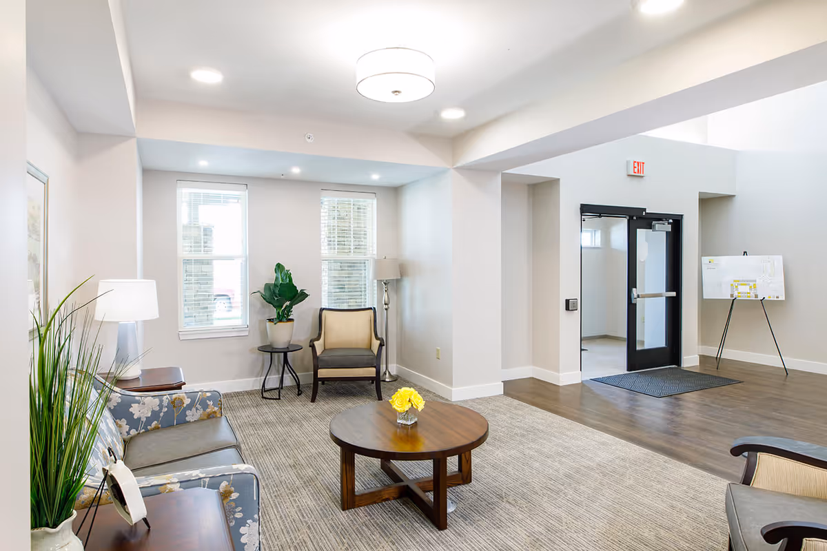 A bright and clean living room area in an assisted living facility featuring a floral patterned sofa, a beige armchair, a round wooden coffee table with a small vase of yellow flowers, two windows with blinds, a floor lamp, and a potted plant. The room has light-colored walls, a carpeted floor, and a doorway leading to another area with a black-framed glass door and an exit sign above it.