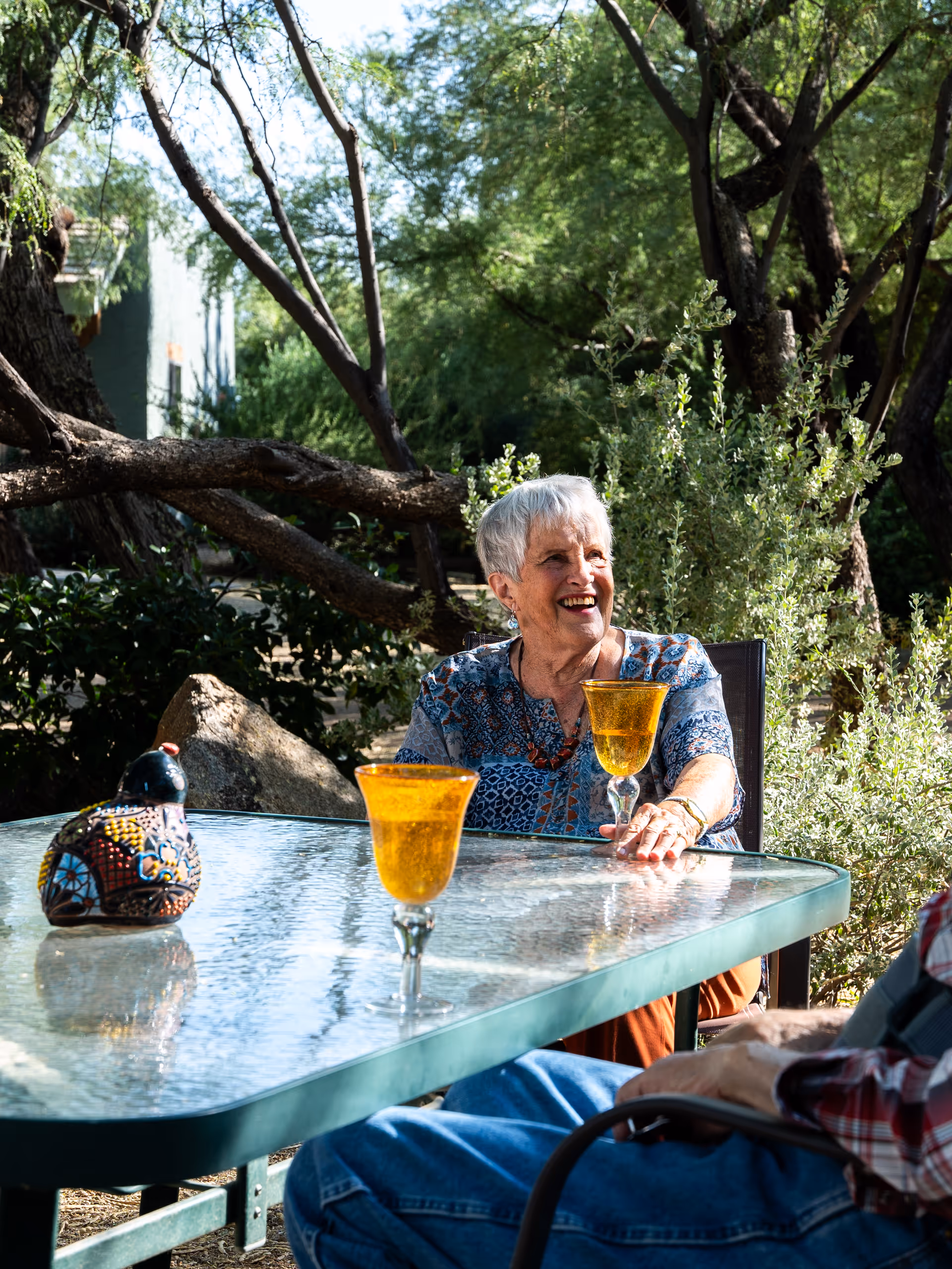 An older woman smiling while seated at a glass outdoor patio table with two amber goblets, surrounded by trees and shrubs.