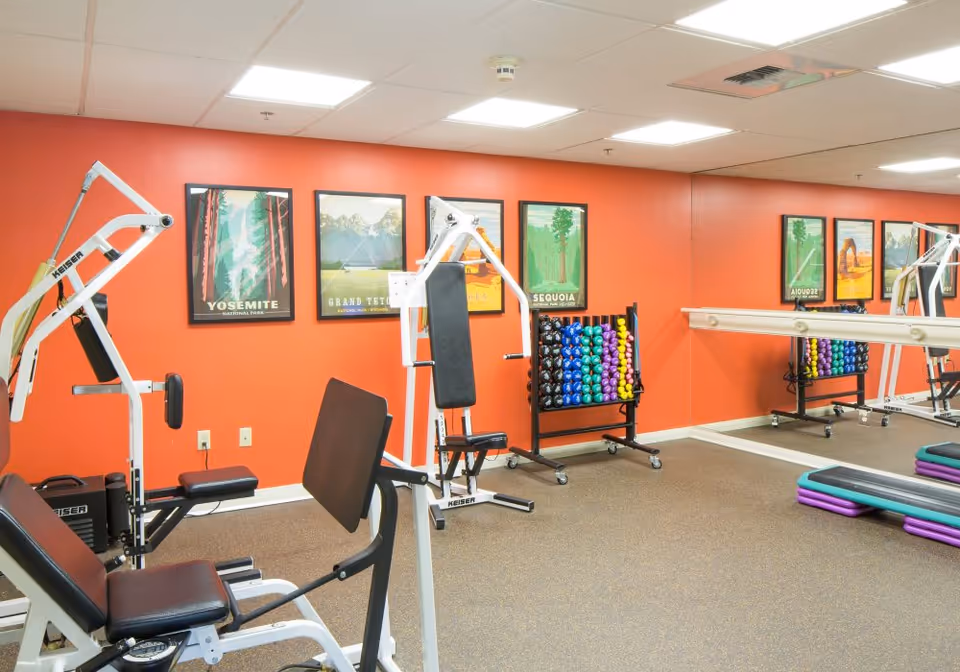 A fitness room with exercise equipment including weight machines, a rack of colorful dumbbells, and step platforms. The walls are painted orange and decorated with framed posters of national parks such as Yosemite, Grand Teton, and Sequoia. A large mirror covers one wall, reflecting the equipment and posters.