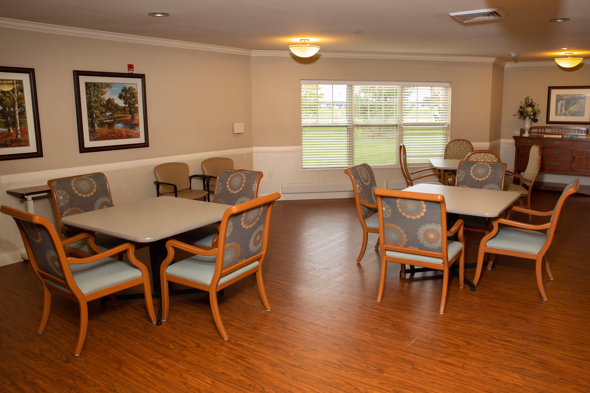 A well-lit dining room with wooden floors, featuring multiple square tables surrounded by cushioned chairs with wooden frames and patterned upholstery. The room has beige walls adorned with framed landscape paintings, a large window with blinds letting in natural light, and a wooden sideboard with a floral arrangement on top.